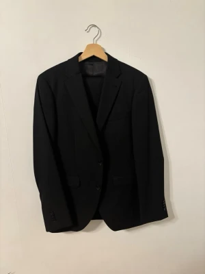 Svart kavaj från Riley Stanton - Säljer en svart kavaj från Riley Stanton med klassisk passform och två knappar framtill. Kavajen har smala slag, innerficka och är helfodrad. Perfekt för dig som vill ha en stilren och tidlös look.