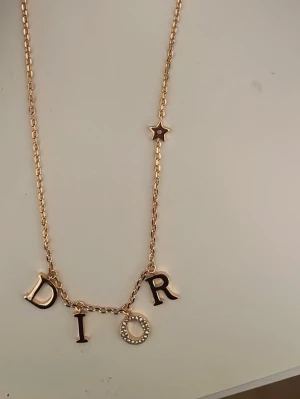 Dior halsband  - Bilderna visar ett halsband från Dior i guldton med bokstäver och en liten stjärndetalj.