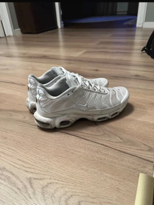 Nike Air Max Plus vita sneakers - Säljer ett par vita Nike Air Max Plus sneakers med mesh- och syntetmaterial, ikoniska Tn-detaljer på hälen och sulan samt synliga luftbubblor. Skorna har snörning och en chunky sula med coola detaljer. Perfekta för dig som gillar streetwear och sportig stil.