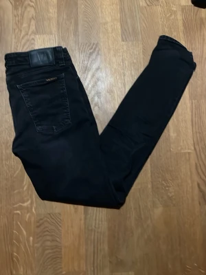 Svarta skinny jeans från Nudie Jeans  - Säljer ett par svarta skinny jeans från Nudie Jeans. De har klassisk femficksdesign, silverfärgade nitar och knapp, samt diskret Nudie-logga på bakfickan. Jeansen är i stretchigt material för extra bekvämlighet och har en smal passform hela vägen ner.
