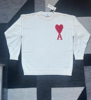 Vit stickad tröja från Ami Paris - Säljer en vit stickad tröja från Ami Paris med röd hjärtdetalj och bokstaven A på bröstet. Tröjan har rund halsringning, ribbade muddar och är långärmad. Perfekt för dig som gillar stilrena plagg med en twist. Helt ny kommer med original förpackningen. Priset kan diskuteras 