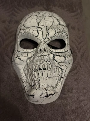 Vit mask med sprucket dödskallemönster - Säljer en riktigt cool mask i form av en dödskalle med sprucket, svartvitt mönster. Masken har svarta detaljer och elastiskt band för att sitta på plats. Perfekt för halloween eller maskerad!