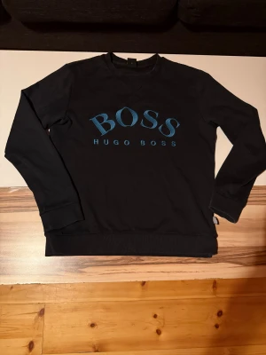 Svart sweatshirt från Hugo Boss - Svart sweatshirt från Hugo Boss med blått BOSS-tryck på bröstet. Tröjan har rund hals, långa ärmar och ribbade muddar vid ärmslut och nederkant. Perfekt för en clean och stilren look.