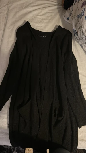 Svart kofta från 157 XS/S - Mysig svart kofta från 157 i storlek XS/S. Koftan är lång, har två fickor framtill och är tillverkad i ett stickat material. Perfekt att slänga på sig över en t-shirt eller hoodie när det är kyligt ute.