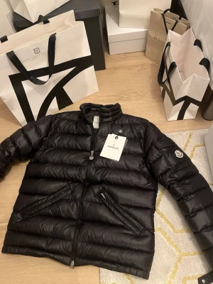 Svart Moncler Agay - Säljer en svart pufferjacka från Moncler med klassisk logga på ärmen och hög krage. Jackan har dragkedja framtill, två snedställda fickor med dragkedja och quiltad design. Materialet är glansig polyester och dunfyllning, perfekt för kyliga dagar.
