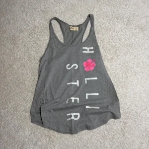 Grå linnetopp från Hollister - Säljer en grå linnetopp från Hollister med vit text och en rosa blomma på framsidan. Toppen har brottarrygg, perfekt för varma dagar eller träning.