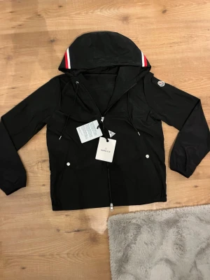 Svart moncler windbreaker ST 2/M - Svart vindjacka från Moncler med huva som har coola röda och vita ränder. Jackan har dragkedja framtill, två fickor med tryckknappar och Moncler-logga på ärmen. Materialet är lätt och känns sportigt, perfekt för dig som gillar stilren design.