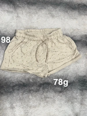 Creamfärgade shorts med hjärtmönster - Säljer ett par creamfärgade shorts med små hjärtan i guld, elastisk midja och snörning framtill. Shortsen har fickor på sidorna, perfekta för varma dagar.🌸 Fint använt skick. Storlek 98