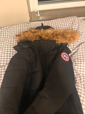Svart Canada Goose dunjacka med päls - Svart dunjacka från Canada Goose med ikonisk röd-vit patch på ärmen och stor huva med fluffig brun päls. Jackan har lång ärm, dragkedja och knappar framtill. Perfekt för kalla dagar och har en clean, klassisk look som alltid är rätt. Jackans dragkedja är sönder men går fixa för typ 100kr man kan använda knapparna annars 