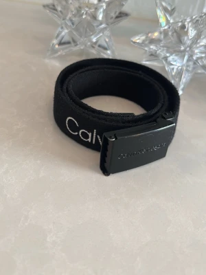 Svart tygbälte från Calvin Klein Jeans - Säljer ett svart tygbälte från Calvin Klein Jeans med vit logotyp längs hela bandet. Bältet har en stilren svart metallspänne med ingraverad logga. Perfekt att styla till jeans eller shorts för en clean look.
