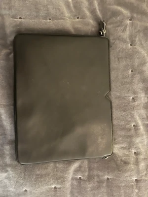 Svart datorväska i skinn med dragkedja - Säljer en stilren svart datorväska i skinn med silverfärgad dragkedja. Väskan har en minimalistisk design och ett rymligt fack på insidan, perfekt för laptop och dokument. Passar dig som vill ha en clean look till plugget eller jobbet.