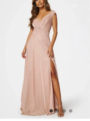 Ljusrosa glittrig långklänning - Supersnygg ljusrosa långklänning med glittrigt tyg, v-ringad omlott upptill och slits på ena sidan. Klänningen har korta ärmar och markerad midja för en elegant siluett. Perfekt för bal eller festliga tillfällen! 