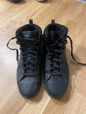 Svarta höga sneakers från Puma - Säljer ett par svarta höga sneakers från Puma med mjukt och fluffigt foder på insidan. Skorna har snörning, rund tå och platt sula. Utsidan är i skinnimitation och de har en klassisk Puma-logga på plösen och sidan. Perfekta för kyliga dagar.
