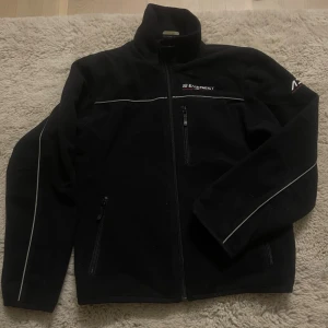Svart fleecejacka från Everest - Svart fleecejacka från Everest med hög krage och dragkedja framtill. Jackan har vita reflexdetaljer längs bröstet och ärmarna, samt tumhål i ärmsluten. Praktiska fickor med dragkedja och broderad logga på bröstet och kragen. Perfekt för kyliga dagar. Storlek 146/152
