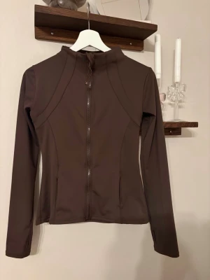 Brun träningsjacka  - Snygg brun träningsjackafigurnära passform och hel dragkedja framtill. Jackan har långa ärmar, hög krage och praktiska fickor. Materialet är mjukt och stretchigt syntet, perfekt för aktiva dagar.