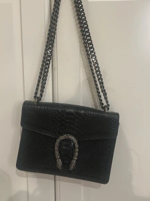 Svart axelväska med kedja och ormdetalj - Snygg svart axelväska med grov kedja som axelrem och cool ormdetalj på framsidan. Väskan har ett reptilmönster i skinn och dragkedja på baksidan samt flera fack inuti. Perfekt för dig som gillar statement-accessoarer.