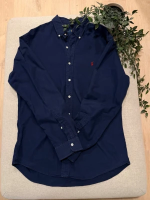 Marinblå skjorta från Ralph Lauren - Snygg marinblå skjorta från Ralph Lauren i slim fit-modell. Skjortan har klassisk krage, långa ärmar och vita knappar framtill. Materialet är mjuk bomull och på bröstet finns Ralph Laurens ikoniska röda logobrodyr.