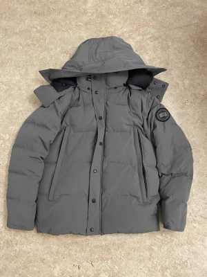 Grå dunjacka från Canada Goose - Säljer en grå dunjacka från Canada Goose med avtagbar huva och svarta detaljer. Jackan har två stora fickor med dragkedja, patch på ärmen och stängs med både dragkedja och knappar. Perfekt för kalla vinterdagar och riktigt snygg streetstil.