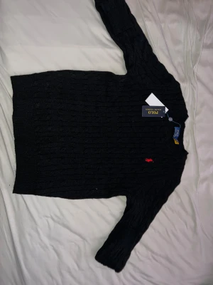 Svart stickad tröja Polo Ralph Lauren - Kabelstickad svart tröja från Polo Ralph Lauren i storlek S. Klassisk modell med rund hals, ribbade muddar och röd broderad logga på bröstet. Perfekt till höst och vinter för en clean och tidlös look.