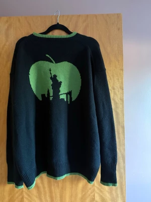 Cloeys tjocktröja - Cool svart stickad tröja med gröna detaljer och motiv av New Yorks skyline och frihetsgudinnan i en grön äpple på ryggen. Texten 'New York' i grönt på ena ärmen. Ribbstickade muddar och krage med grön kant. Perfekt för dig som gillar streetstyle och unika prints. Tjocktröja i kashmir/ullmix från Chloe Schuterman, city intarsia New York sweater. Finns inte till försäljning längre. Onesize. Nypris ca 6000kr. 🍏