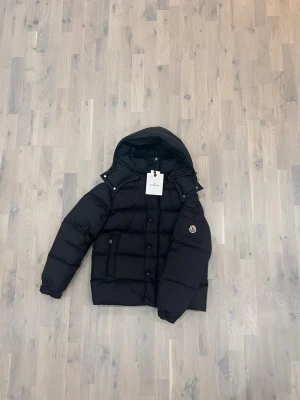 Svart Moncler pufferjacka med huva - Svart pufferjacka från Moncler med huva och klassisk logga på ärmen. Jackan har quiltad design, dragkedja och knappar framtill samt praktiska fickor. Insidan har ett coolt serietidningsmotiv. Perfekt för dig som vill ha en varm och snygg jacka i vinter.
