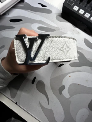 Vitt L v bälte 115cm - Säljer ett stilrent  bälte i vitt skinn med klassisk logga i silver framtill. Bältet har diskret monogrammönster och är tillverkat i Spanien. Perfekt accessoar för att lyfta din outfit med exklusiv känsla.