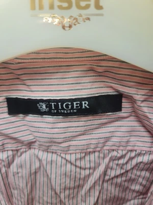 Randig skjorta från Tiger of Sweden - Säljer en snygg skjorta från Tiger of Sweden med smala rosa och vita ränder. Skjortan har klassisk krage, långa ärmar och knappar framtill. Materialet är mjuk bomull som känns skönt mot huden. Perfekt för dig som gillar stilrena och fräscha plagg.