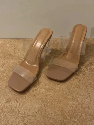 Beige klackpumps med transparent rem - Stilrena pumps i beige med transparenta remmar och fyrkantig tå. Skorna har en hög, genomskinlig klack och platt sula framtill. Perfekta för dig som vill ha en modern och trendig look.