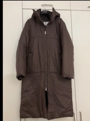 Brun lång pufferjacka Tiger of Sweden - Helt ny lång, brun pufferjacka med huva och raka linjer. Jackan har totalt fyra fickor framtill, dragkedja hela vägen och bekväma muddar detaljer vid ärmsluten, även samma mudd material vid kragen vilket är super bekvämt och varmt! Perfekt för kalla dagar och ger en clean, oversized vibe då den är lite större i storleken☺️Nypris:799euro/87050sek!