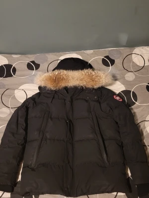 Svart Canada Goose dunjacka med päls - Säljer en svart Canada Goose dunjacka med fluffig päls på huvan och klassisk logga på ärmen. Jackan har lång ärm, dragkedja och knappar framtill. Perfekt för kalla dagar med sin tjocka och quiltade design. Huvan är stor och ger extra skydd mot vind. XL men passar L