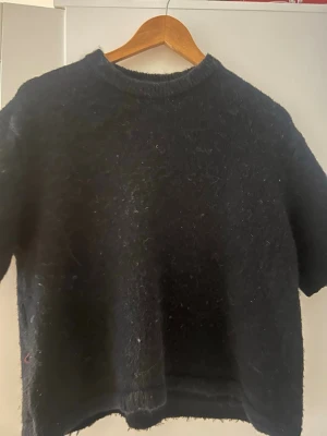 Svart stickad tröja från Gina Tricot - Mysig svart stickad tröja från Gina Tricot i storlek XS. Tröjan har korta ärmar, rund hals och en lite fluffig yta som ger en cool look. Perfekt för lager-på-lager och passar till både jeans och kjol. Enkel och stilren design.