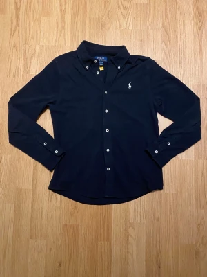 Mörkblå skjorta från Polo Ralph Lauren - Snygg mörkblå skjorta från Polo Ralph Lauren med klassisk krage och vita knappar. Broderad logga i vitt på bröstet. Skjortan är långärmad och har en stilren, normal passform. Tillverkad i mjuk bomull som känns skön mot huden. Storlek 14-16 som motsvarar Xs. Skjortan är knappt använd och har inga defekter. Hör av dig om du har mer frågor. Priset är prutbart via enkel affär.