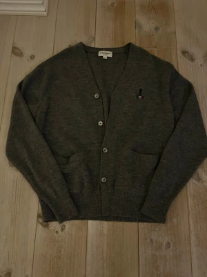 Grå cardigan från Yves Saint Laurent - Snygg grå cardigan från Yves Saint Laurent i storlek M med broderad logga och Paris-detalj på bröstet. Materialet är mjukt och stickat, perfekt för lager-på-lager under kyliga dagar. Fraktar på 1-2 dagar!