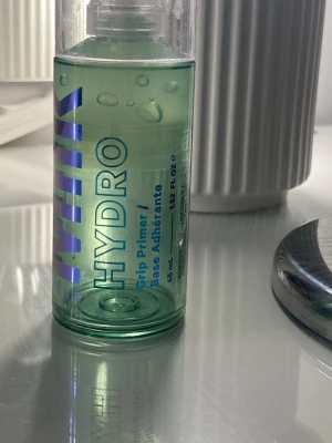 Milk Hydro Grip Primer 45 ml - Säljer en Milk Hydro Grip Primer i genomskinlig grön plastflaska med pump. Primern är gelbaserad och ger en återfuktande känsla. Perfekt för att få sminket att sitta längre och ge en fräsch finish. Flaskan rymmer 45 ml och har blå och lila text på framsidan.