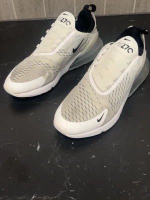 Nike Air Max 270 vita sneakers - Nike Air Max 270 sneakers i vit och grå färg med svarta detaljer. Skorna har en stor synlig Air-enhet i hälen, mesh-ovandel för bra ventilation och en platt sula. Snygg design med logga på tungan och sidan, samt snörning och dragflik bak.