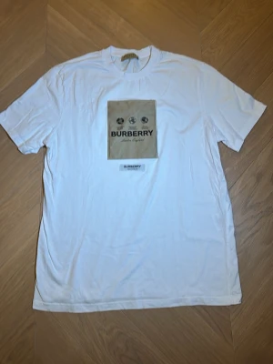 Vit Burberry t-shirt med beige patch - Säljer en vit t-shirt från Burberry med en stor beige patch på bröstet och svart Burberry-tryck. T-shirten har rund hals och korta ärmar. Materialet är mjuk bomull och den har en clean, klassisk vibe med tydlig logga framtill.