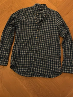 Blårutig långärmad skjorta från Gant - Säljer en blårutig skjorta från Gant med klassisk krage och knappar framtill. Skjortan är tillverkad i bomullspoplin och har en snygg, tidlös design med ljusa och mörka blå toner. Perfekt för dig som gillar stilrena och bekväma skjortor med lite preppy känsla.