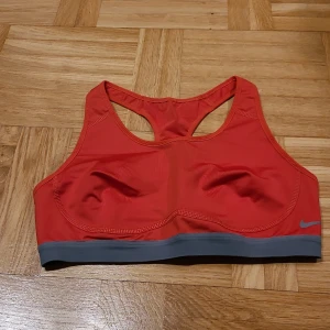 Röd sporttopp från Nike Dri-Fit - Nike Dri-Fit sporttopp i rött med grå kant nedtill. Modellen är ärmlös med brottarrygg och har en liten Nike-logga på framsidan. Tillverkad i syntetmaterial som andas och håller dig torr under träningen. Perfekt för gymmet eller löprundan.