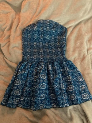 Blå bandeau kortklänning med mönster - Supersöt blå bandeau-klänning med vitt paisleyinspirerat mönster. Klänningen är kort och har en smockad överdel som sitter tajt över bröstet och en utställd kjol. Perfekt för varma dagar och ger en chill sommarvibe.