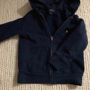 Mörkblå huvtröja från Polo Ralph Lauren med Zip - Säljer en mörkblå huvtröja från Polo Ralph Lauren med dragkedja och en broderad gul logotyp på bröstet. Tröjan har långa ärmar och en praktisk huva. Perfekt för en avslappnad stil. 🧢