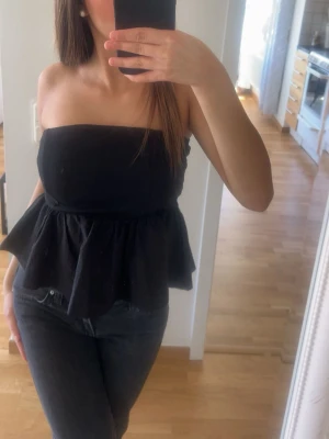 Svart bandeau tubtopp med volang - Säljer en svart bandeau tubtopp med volangkant nedtill. Toppen är axelbandslös och har en snygg, croppad passform som ger en trendig siluett. Perfekt att matcha med jeans eller kjol för en cool look.