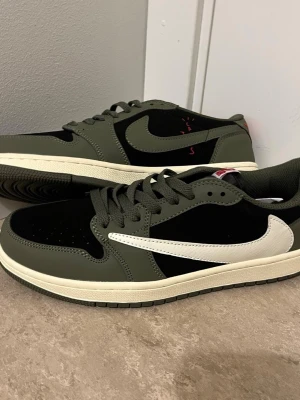 Nike Air Jordan 1 Low Travis Scott - Nike Air Jordan 1 Low Travis Scott i olivgrön och svart med vit omvänd swoosh. Skorna har röda broderade detaljer på hälen och klassisk snörning. Ovandelen är i skinn och mocka, med en platt sula och rund tå. Perfekt för dig som gillar streetwear och sneakers med unik design.