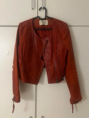Röd skinnjacka från ONLY - Säljer en röd skinnjacka i äkta skinn från ONLY med cool omlott-design och dragkedjor både fram och vid ärmsluten. Jackan är croppad.