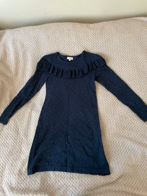 Marinblå barnklänning med volang Pomp de Lux - Snygg marinblå barnklänning från Pomp de Lux med lång ärm och volangdetalj över bröstet. Klänningen har små prickar över hela tyget och är i mjuk bomull. Perfekt för dig som gillar stilrena detaljer och bekväma material.