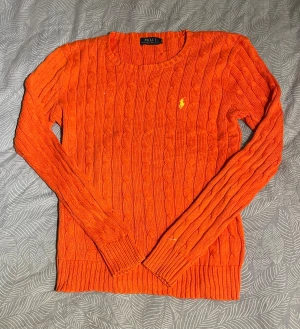 Orange stickad tröja från Polo Ralph Lauren - Säljer en snygg orange kabelstickad tröja från Polo Ralph Lauren med rund hals och lång ärm. Tröjan har det klassiska Polo-logot broderat i gult på bröstet och ribbade muddar vid ärmslut och nederkant. Perfekt för dig som gillar färg och stil!