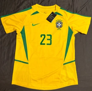 VM 2002 Brasilien tröja  - Säljer en riktigt fin Brasilien VM 2002 tröja med Kaká 23. Tröjan är helt ny och i perfekt skick, inga defekter eller slitage. Färgerna är klara, trycket sitter perfekt och materialet känns fräscht och bra. Passar både samlare och dig som vill ha en ikonisk Brasilien-tröja med en av landets största spelare genom tiderna.