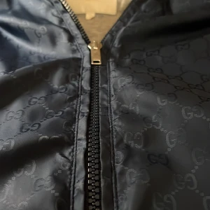 Mörkblå väst från Gucci med GG-mönster - Gucci påsen som man kan lägga in västen. Snygg mörkblå väst från Gucci med ikoniskt GG-mönster över hela tyget. Västen har dragkedja framtill, ståkrage och grått innerfoder med en praktisk innerficka. Perfekt för dig som vill ha en exklusiv och stilren look. Finns en luva att fälla ut! Den är för tolv och 13 år, en jävligt fet väst. Har påse till om man vill ha den är och Gucci!