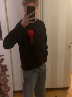 Svart stickad tröja från Ami Paris - Svart stickad tröja från Ami Paris med röd hjärtlogga och bokstaven A på bröstet. Tröjan har rund halsringning, ribbade muddar och en klassisk passform. Perfekt för dig som vill ha en stilren men ändå unik look.