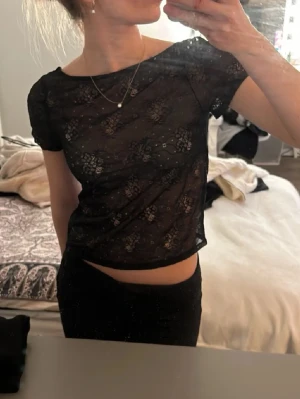 Svart spetsblus med blommönster - Säljer en svart, transparent blus med korta ärmar och blommigt spetsmönster. Toppen har en rundad halsringning, djup rygg och är croppad i modellen. Perfekt att styla med ett linne under eller som den är för en cool look.