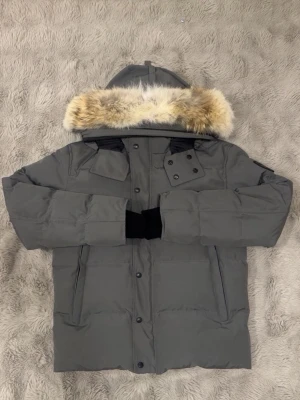 Grå dunjacka från Canada Goose - Säljer en grå dunjacka från Canada Goose med fluffig päls på huvan och snygga svarta detaljer. Jackan har lång ärm, flera fickor och knappar framtill. Perfekt för kalla vinterdagar och har en stilren look med märkets patch på ärmen.